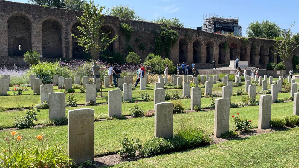 An open day at Rome War Cemetery. Image shows visitors being given a guided tour around the cemetery. Several lines of headstones are visible, wiht the Stone of Remembrance in the top right. The Aurellian Walls of Rome run along the back of the cemetery.