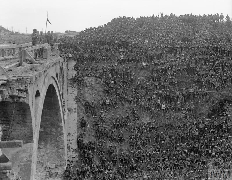 Thousands of British infantrymen sit on a railway cutting listening to a speech by a British General. The General and staff are standing on a thick stone bridge whose arches span a canal.