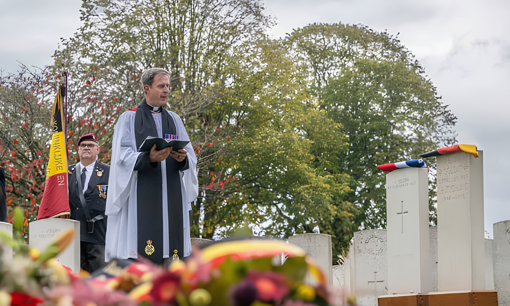 A military padre reads from the bible next to the grave site of a British and German soldier. British and German headstones mark the grave, with the repsective nations' flags placed atop each. A soldier wearing a beret and holding a Belgian flag can be seen behind the priest.