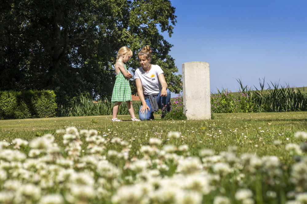 A blonde girl in a green dress stands next to her kneeling mother beside a CWGC headstone.