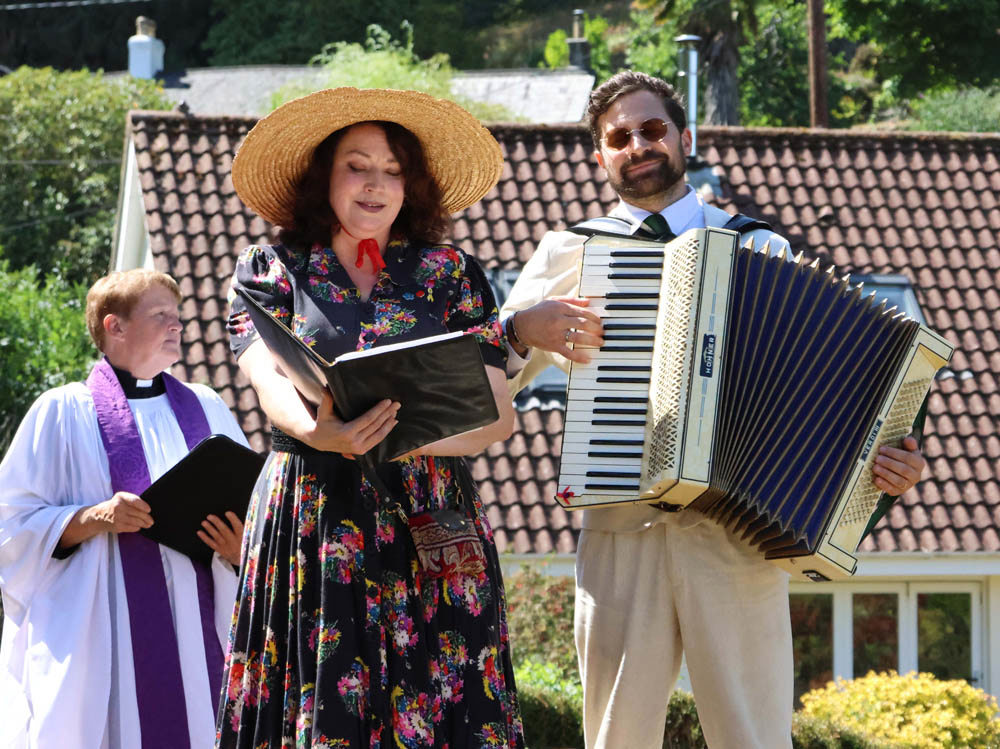 Women in a sundress and strat hat reads from a book while a man in a cream suit plays an accordion.