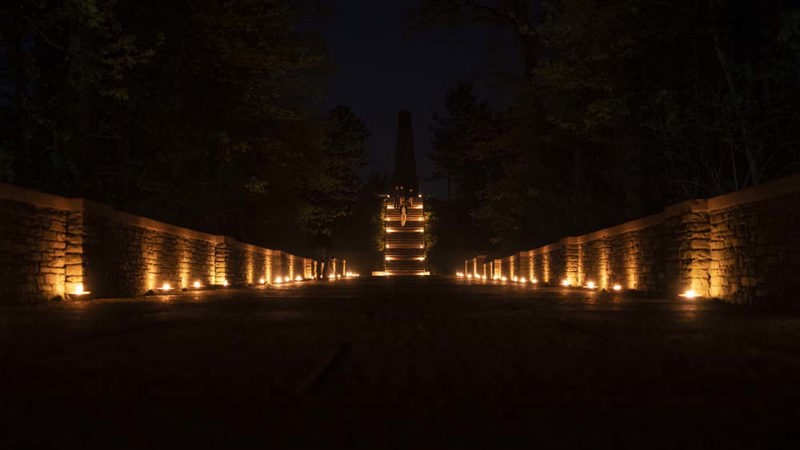 Buttes New British Cemetery central pathway at night illuminated with a warm golden glow by rows of candles.