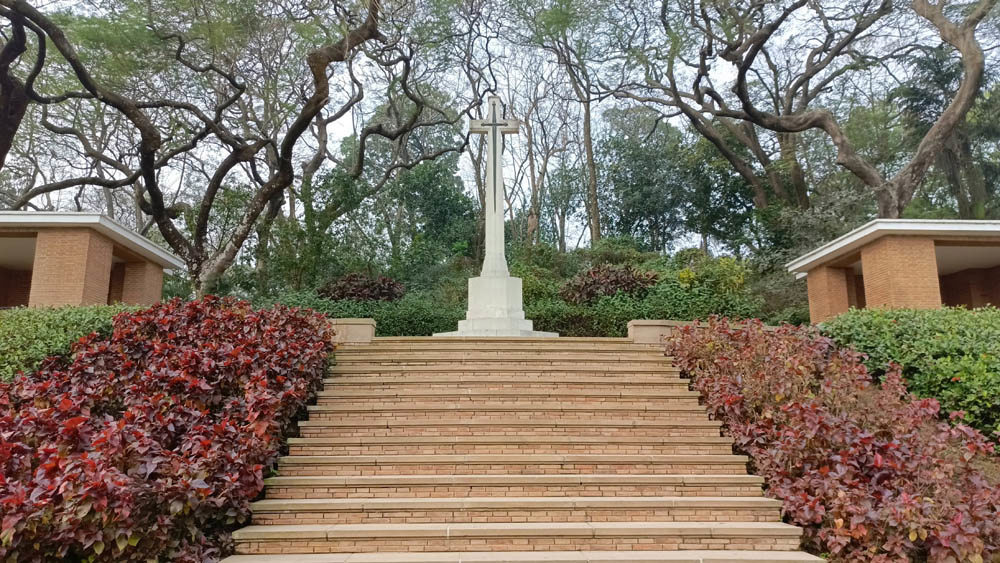 Cross of sacrifice atop a flight of redstone steps in Maynamati War Cemetery. A bush with red flowering plants and a green leafy bush flank the staircase.