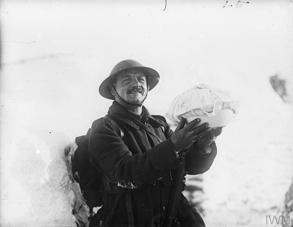 A British soldier gleefully holds up his wrapped Christmas pudding on the Western Front. The background and trench is covered in deep white snow, while the solder is wearing a tin helmet and winter great coat, leaning against the snowy trench.