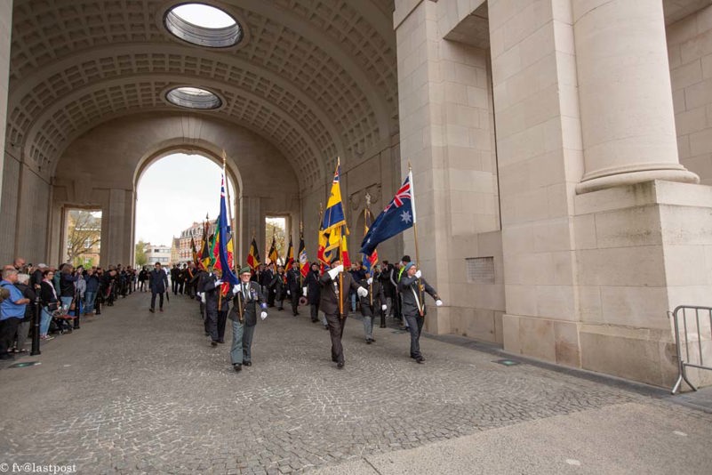 A group of Australian and New Zealand Veterans parade through the stone arch of the Ypres (Menin Gate) Memorial, each holding their nation's respective flag, at the head of a ceremonial column,