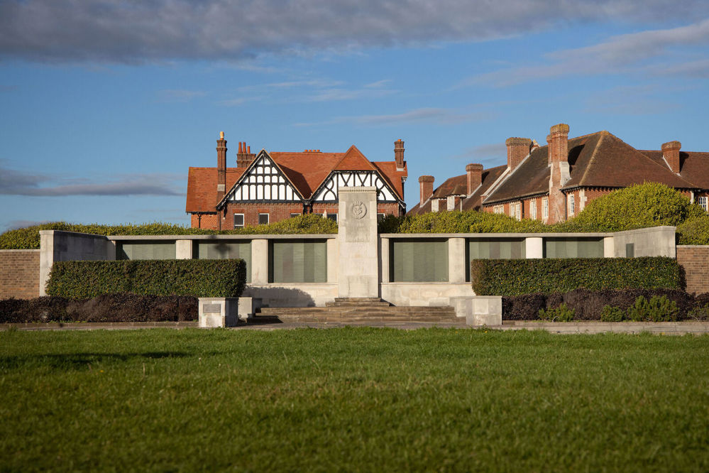 Lee-on-Solent Naval Memorial showin the memorial's aged bronze name panels set on two wings either side of the central rectangular memorial. A low hedge leading up to the stone memorial is visible set before a green lawn. A mock-tudor, red-roofed house is visible behind the memoral.