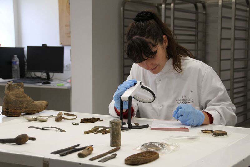 A woman in a white lab coat and blue gloves examines WW1-era artefacts using a microscope