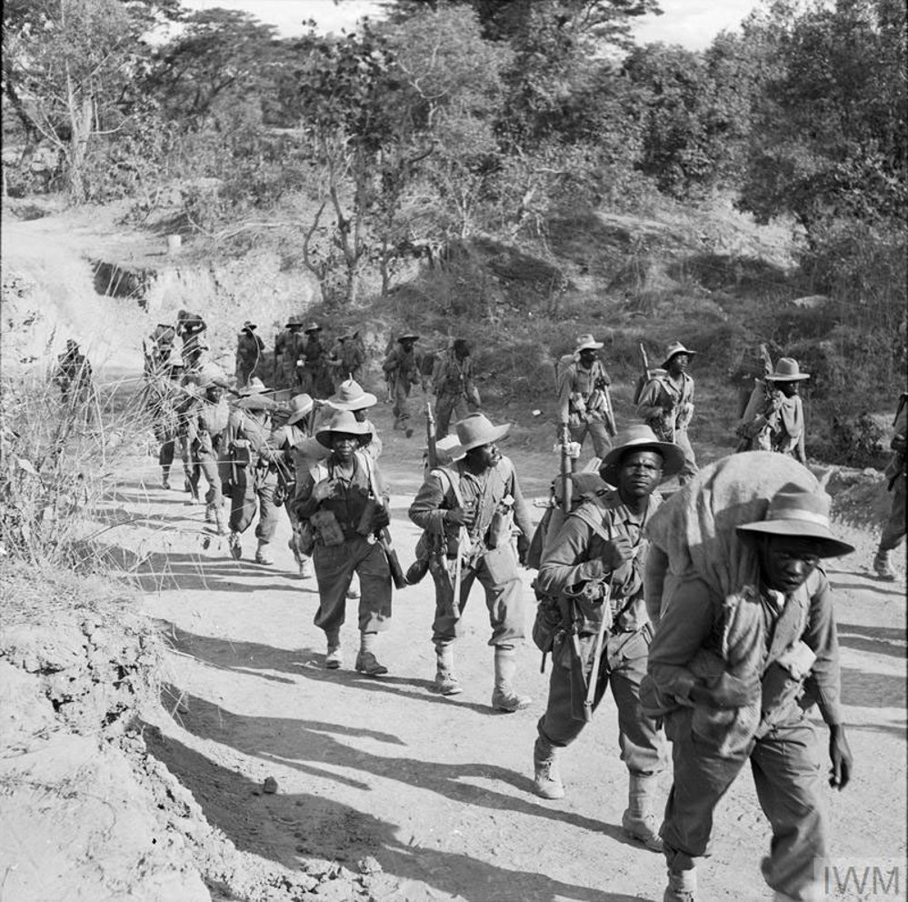 East African soldiers carrying packs and rifles march up a road through tree-covered hills in Burma.