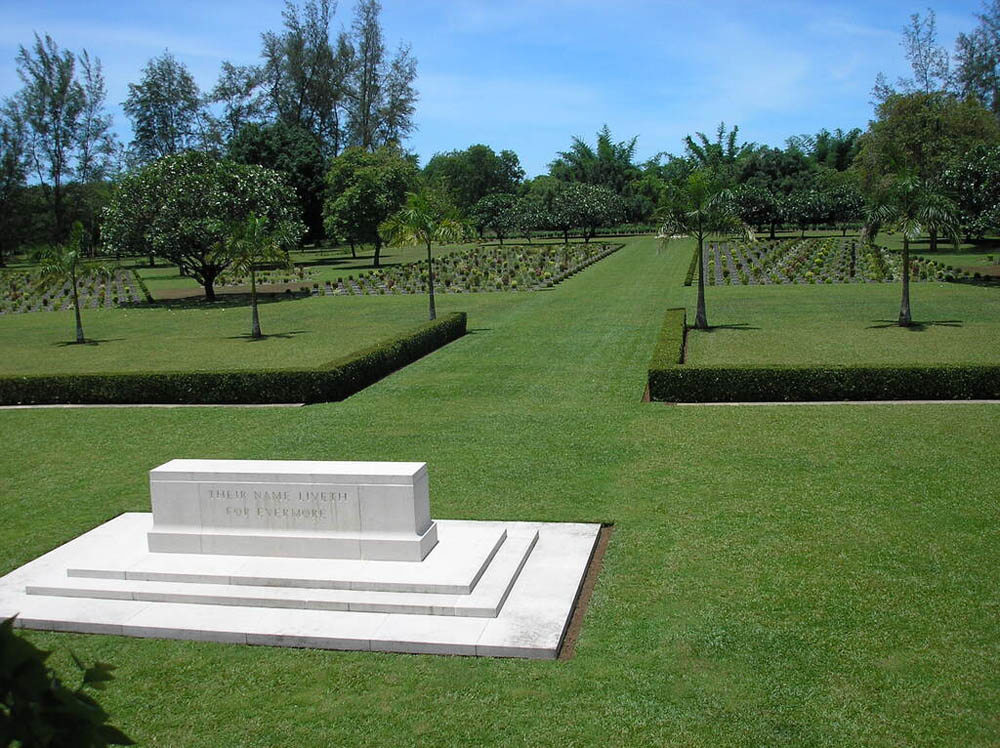 Clean white stone Stone of Remembrance sits on a wide, well-kept green lawn at Thanbyuzayat War Cemetery. Two low hedges flank a pathway towards rows of CWGC headstones. Trees dot the area.