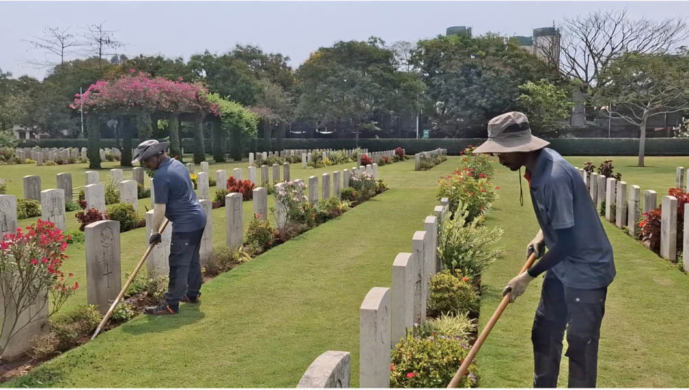 Two CWGC gardeners use long roes to cultivate the plant borders at Madras War Cemetery. Along headstone rows next to the workers you can see a variety of blooming, healthy local plants.