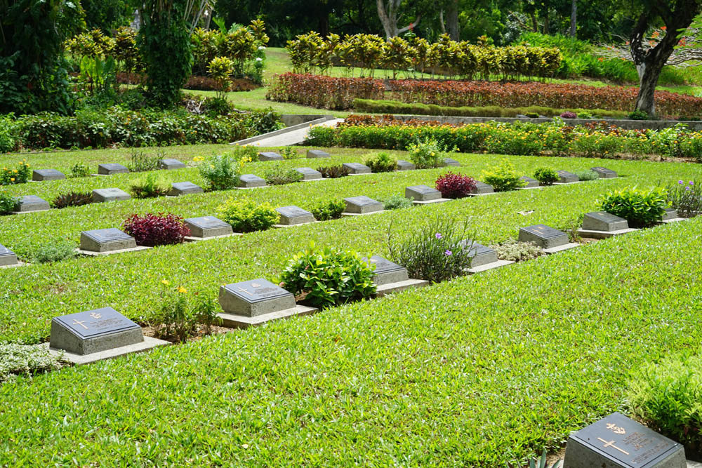 Rows of bronze headstone plaques at Ambon War Cemetery set amid blooming plant borders and a lush green lawn.