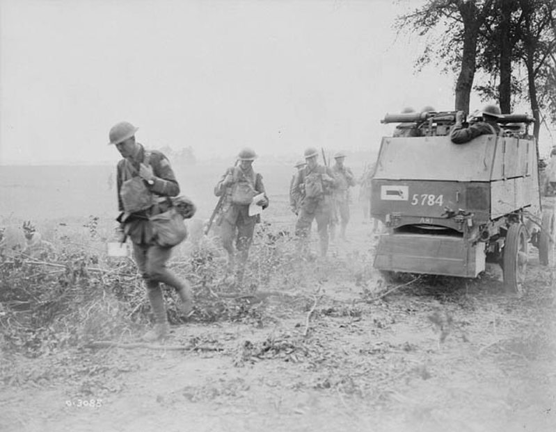 Canadian soldiers on the move along a dusty road, supported by a boxy Great War Autocar. A thin row of trees is visible in the background.