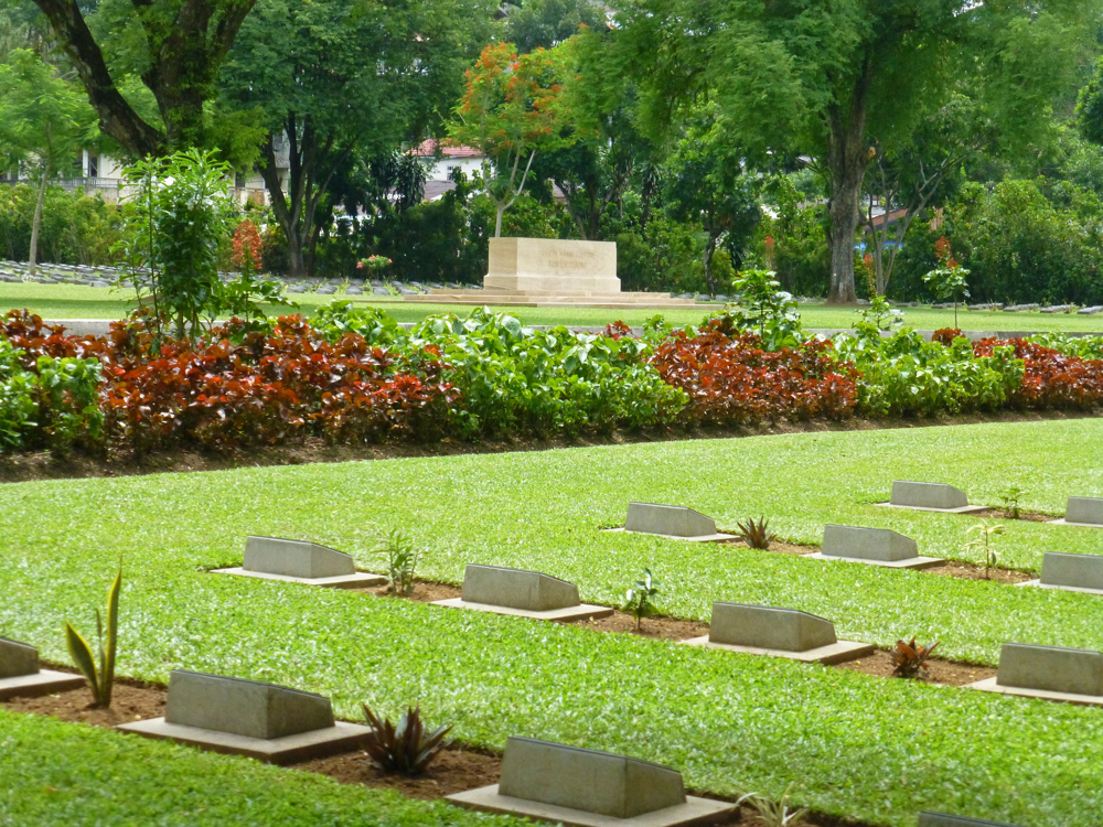 Bronze grave markets peaking out of their burial plot on a shimmering emerald lawn at Ambon War Cemetery. A series of red and green bushes are visible in a planter running alongside the lawn. In the background, the altar like CWGC Stone of Remembrance can be seen.