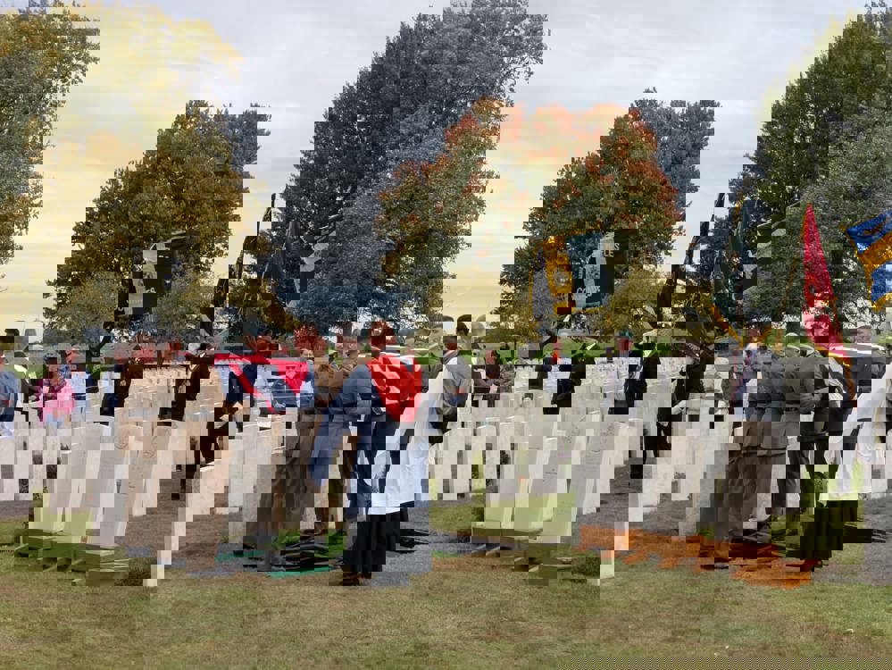 British soldiers lower a union-jack shaped coffin into a war grave in Warlencourt British Cemetery. A series of CWGC headstones waiting installation on wooden posts are visible to the right. In the background, standard bearers hold various military banners.