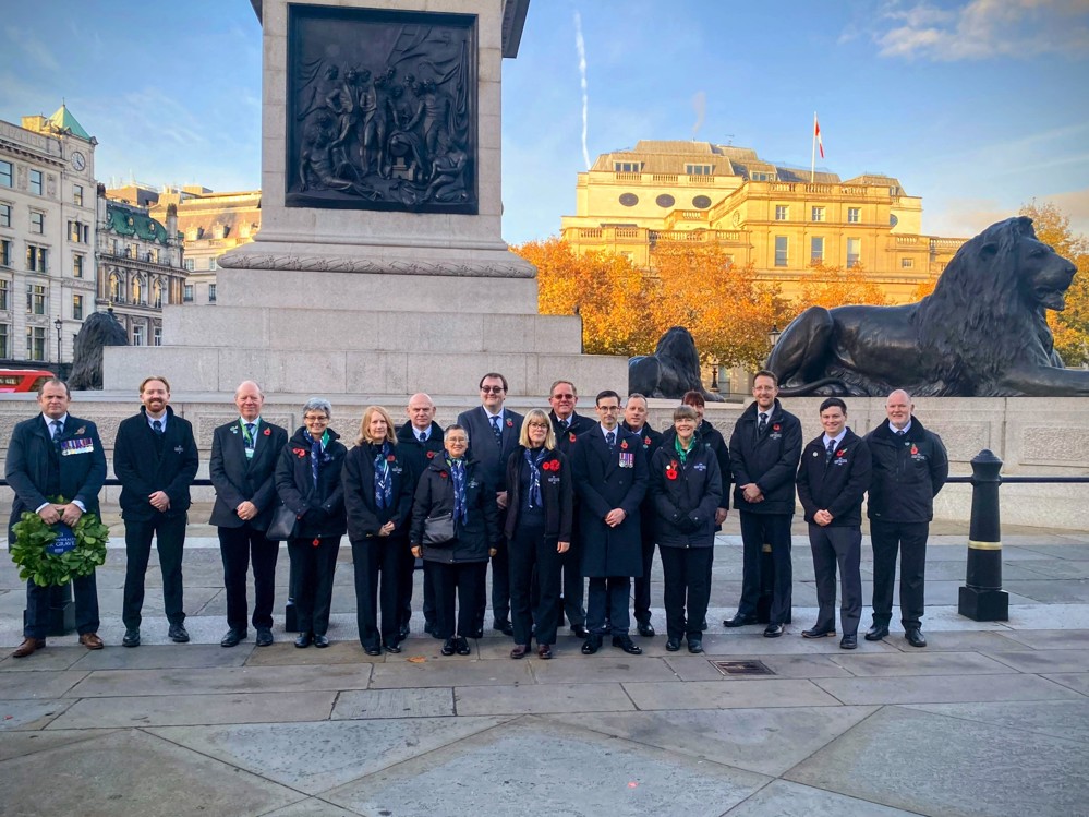 CWGC Team poses at the base of Nelson's Column, in London, for Remembrance Day Ceremonies. Each staff member is wearing dark clothing, punctuated by bright red poppy badges pinned to their outfits.