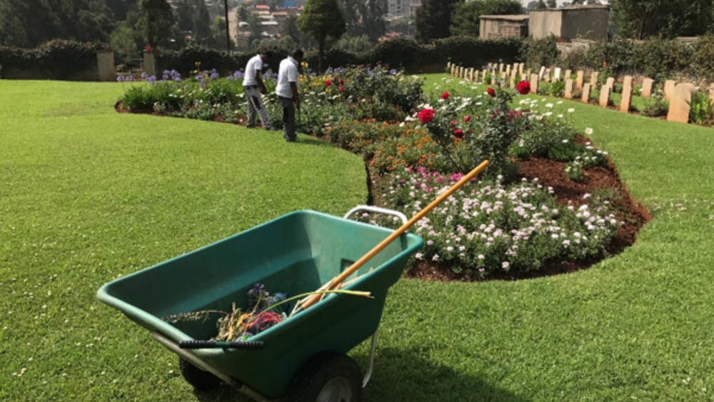Gardeners attending to a large planter full of colourful plantlife, including red roses, smaller pink flowers, white daisies and purple headed varieties. In the foreground is a wheelbarrow with a protruding spade handle.
