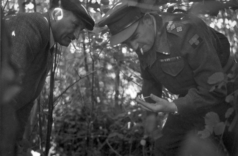 A British officer inspects an ID disc of a fallen soldier alongside a French civilian.