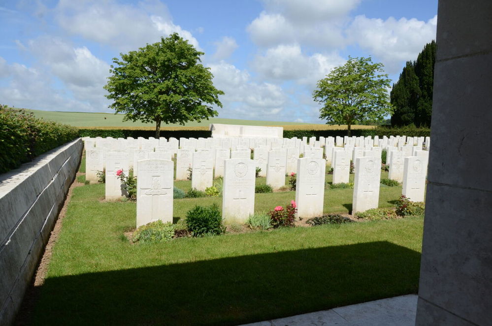 Headstones, rear boundary wall, and Stone of Remembrance as seen from one of the shelter buildings at Forceville cemetery. Two large verdant trees are visible behind the cemetery, as are lush rolling green fields set under a clouded blue sky.