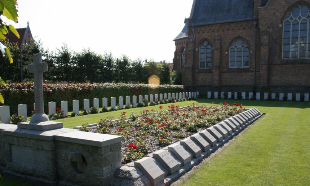 German war graves plot at Sint Donaas Churchyard, Zeeburgge, Belgium. The German graves are in a single plot planted with red roses, with a stone border. Carved into the border are grave markers naming the men buried here. A stone cross atop an altar-like bondary caps the plot. Around the lawn is a ring of CWGC headstones.