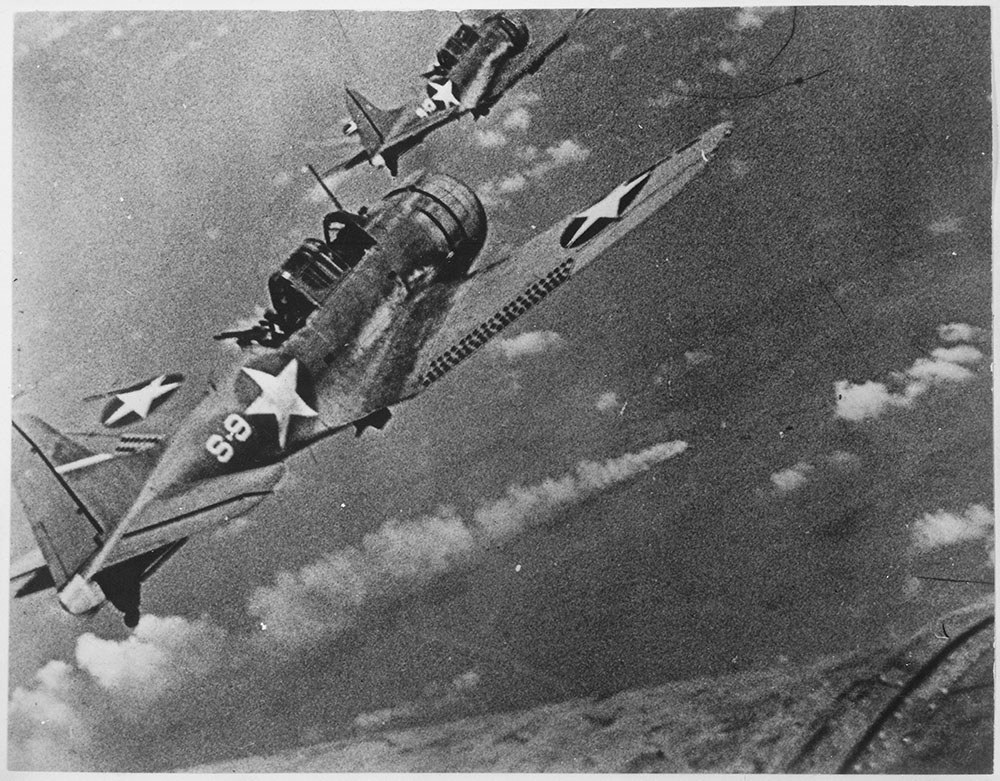 Two US navay aircraft flying high over the Battle of Midway. Way below, puffs of white clouds and grey smoke emanating from damaged ships waft skywards.