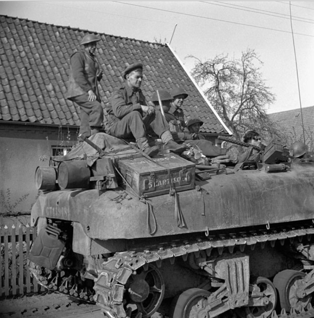 A group of Canadian soldiers riding on the back of a Sherman tank past a low, sloped-roofed building and a skeletal tree on the way into Germany. Lots of equipment and storage boxes have been stowed atop the tank.