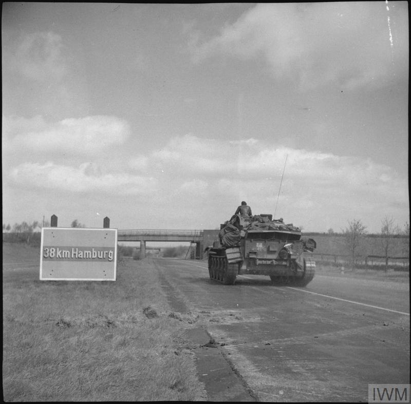 A British Second World War Cromwell tank driving along a German autobahn. A sign says "38 km Hamburg" in Gothic font.