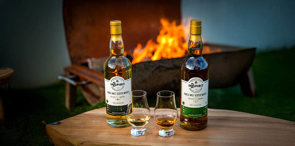 Two bottles of whiskey with glasses holding a small dram sit on a wooden table in front of an open half-drum barbeque. A bright flame is burining on the barbeque.