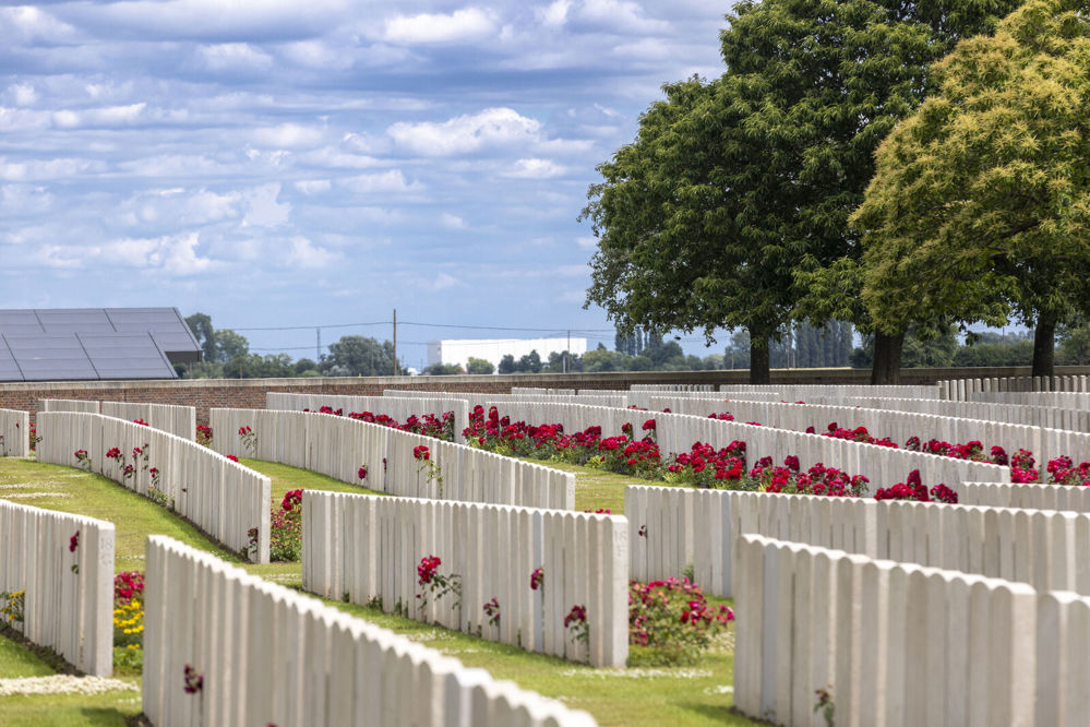 Rows of white, round topped CWGC headstones in Lijssenthoek Military Cemetery. Red blooming roses have been planted in some of the grave plot borders. Tall trees are visible in the background. The sky is a light blue dappled with fluffy white clouds.