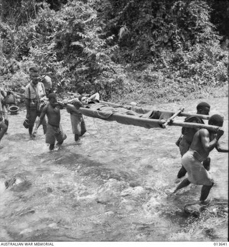 Native Papuan stretcher bearers carry a wounded Australian soldier over a swift coursing stream in the middle of a jungle.