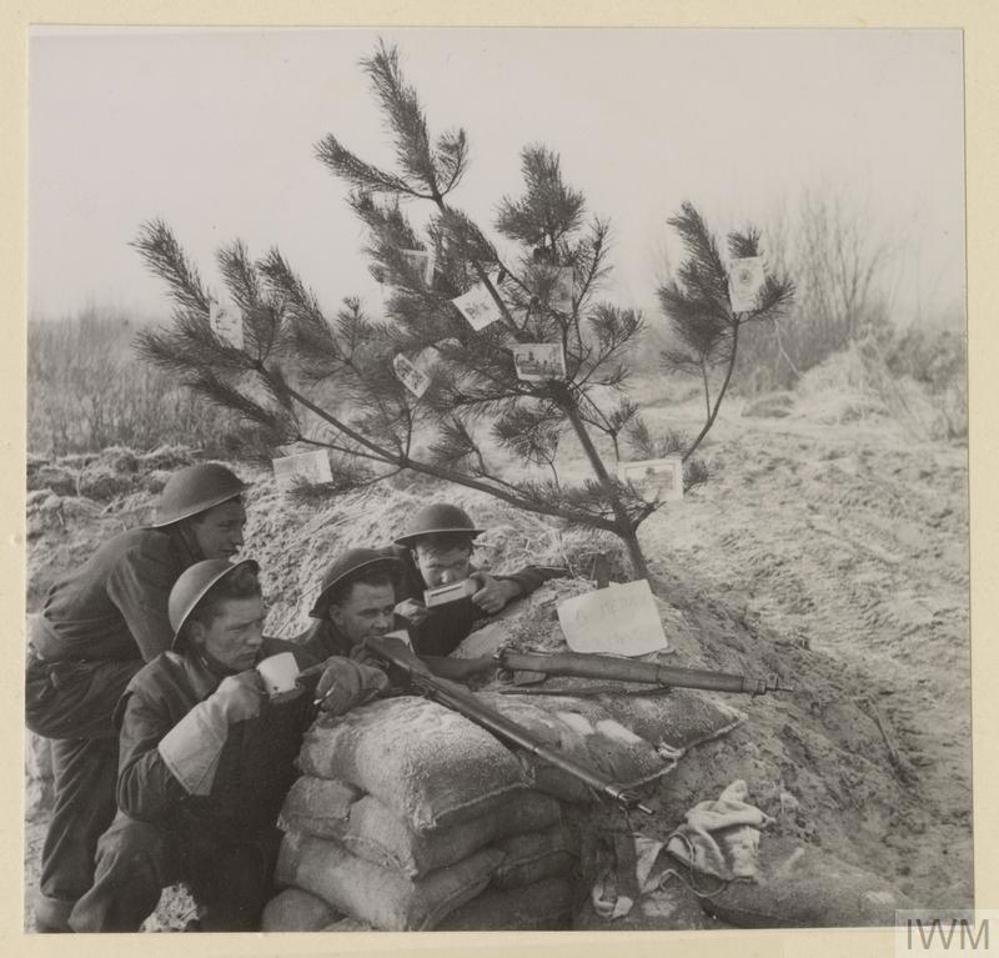 British soldiers rest on sandbacks with their rilfes, while drinking tea, under a thin tree decorated with Christmas Cards.