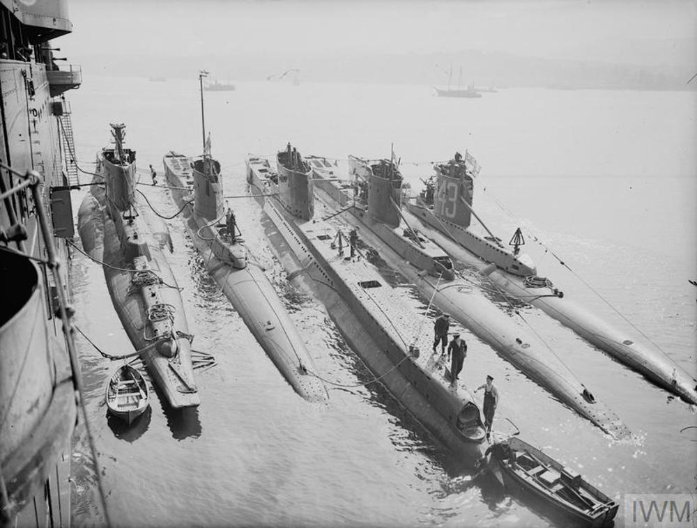 Submarines of various classes and sizes berthed at Holy Loch. Crewmen can be seen working on the vessels, while a series of small boats are bobbing alongside the submarines.