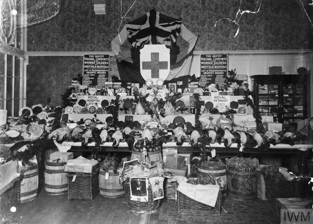 A large pile of christmas gifts, foodtsuffs, and puddings arrayed on a table, ready for delivery to British troops during the First World War. Several barrels and crates sit beneath the table. A backdrop of a Union Jack and a large Red Cross shield sits at the back of the table.