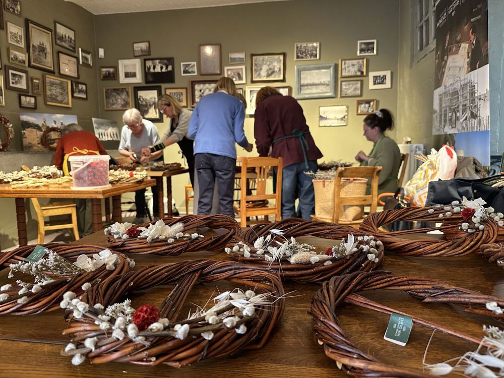 Close up of wreaths on a table. In the background, you can see a group of people crafting wreaths with their hands. Supplies kept in transparent boxes are visible on the workshop tables.