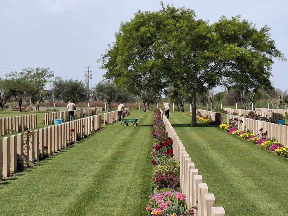 Gardeners hard at work caring for the grave plot borders at Catania War Cemetery. A variety of pink, purple, red, and maroon flowers bloom from the flower beds, contrasting with the creamy white CWGC headstones. A large tree shades one of the headstone rows.