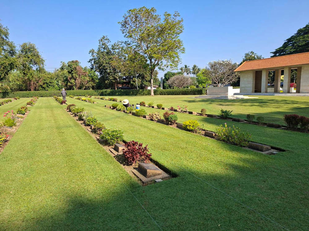 View of Chungkai War Cemetery showing a worker working in the background, wide, neatly kept lawns, and bronze grave markers set well kept borders.