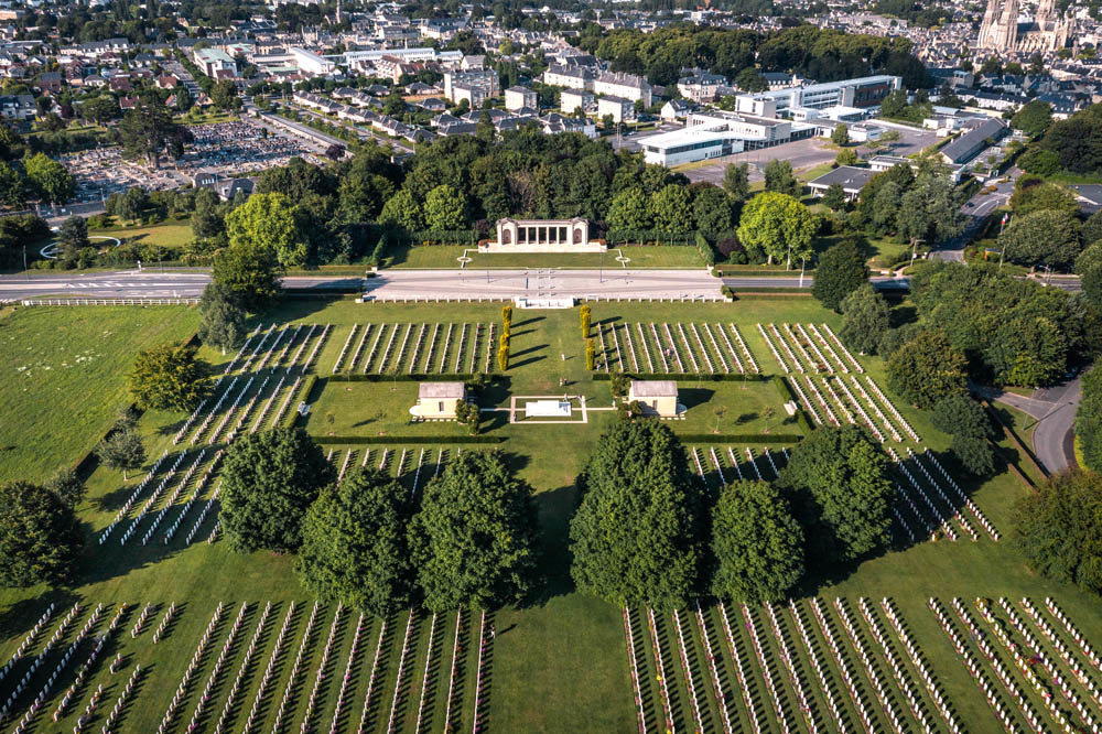 Aerial view of Bayeux War Cemetery and Memorial showing rows of headstones, tree planted avenues, and the Stone of Remembrance in the cente of the cemetery. Set to the back of the site, across a road bisecting the cemetery, sits the Roman-esque Bayeux Memorial.