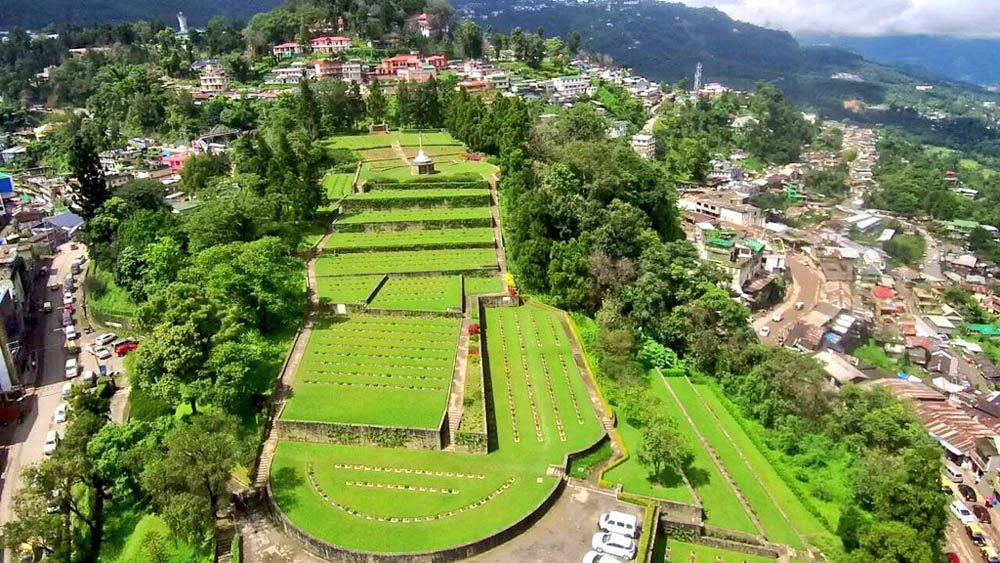 Aerial view of Kohima War Cemetery showing its terraces, plots, and war graves. The cemetery sits atop a hill, flanked by city buildings and roads. Trees ring the summit of the hill.