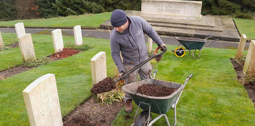 A nab shovels compost out of a wheelbarrow into the plant border of a CWGC grave plot.