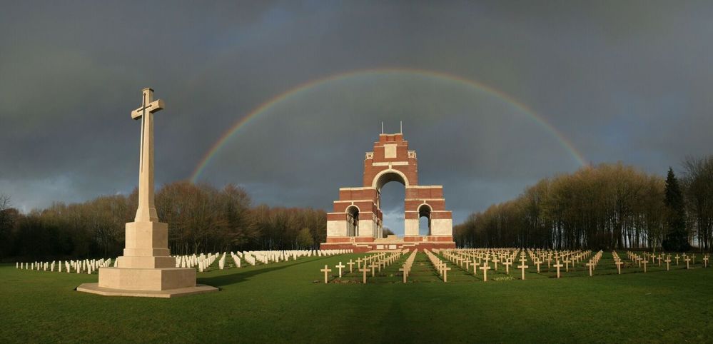 Thiepval Memorial as seen from Thiepval Anglo-French Cemetery. The Cross of Sacrifice is visible in the foreground. A rainbow arcs over the memorial in the background.