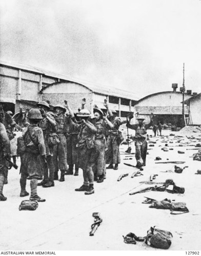 British soldiers raise their hands in surrender to Japanese soldiers during the fall of Singapore. Discarded rifles, packs, helmets, and equipment litter the floor.