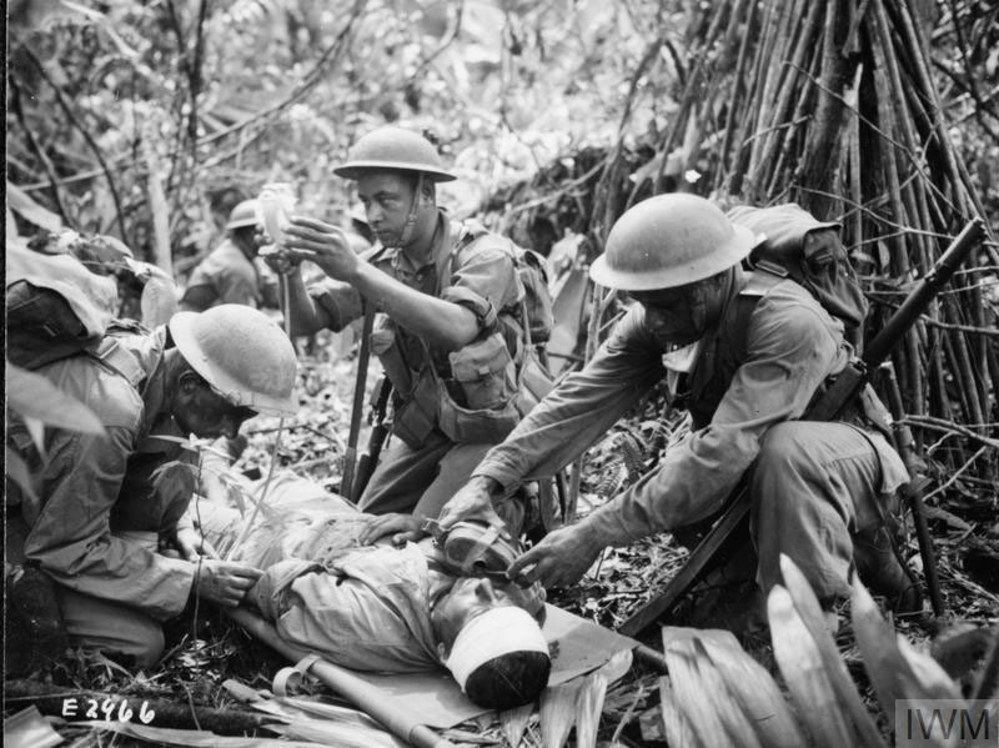 A Fijian medical orderly administers an emergency plasma transfusion to a wounded soldier on a stretcher in a jungle during WW2.
