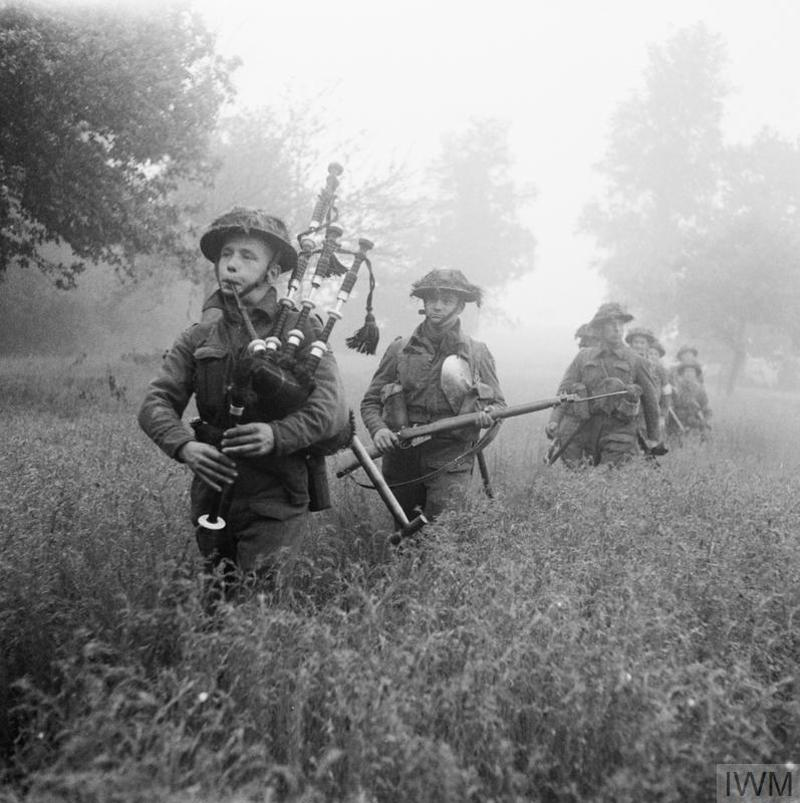Scottish WW2 era infantry advance through long grass and mist during Operation Epsom. The lead soldier is playing the bagpipes.