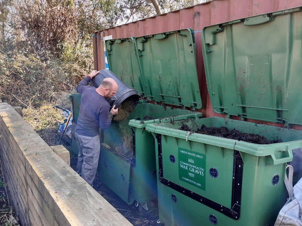A man throws gardening waste into a large green plastic compost bin. The bin is marked with a Commonwealth War Graves Commission sign.