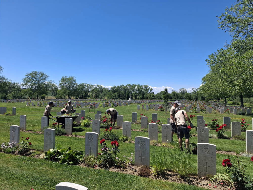 CWGC gardeners in light taupe polo shirts and shorts working on the lawn and plant borders at Coriano Ridge War Cemetery.