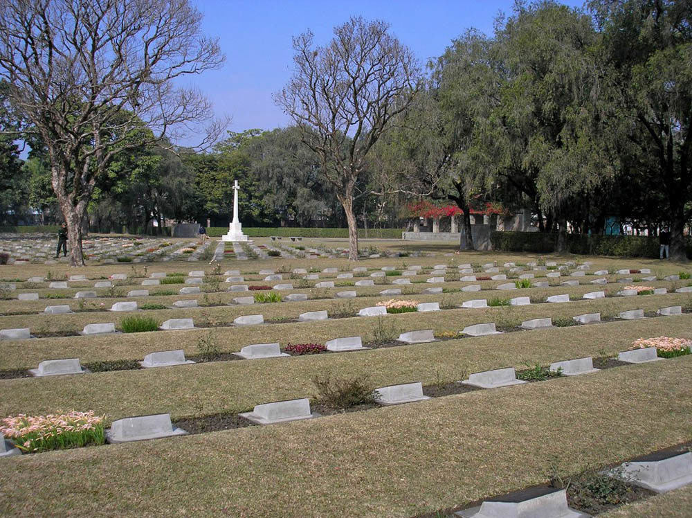 Stone, bronze-faced war markers arrayed in plots and rows at Imphal War Cemetery. The grass is dry and turning from green to a more hay-like colour. Trees and a white stone Cross of Sacrifice at Imphal War Cemetery.
