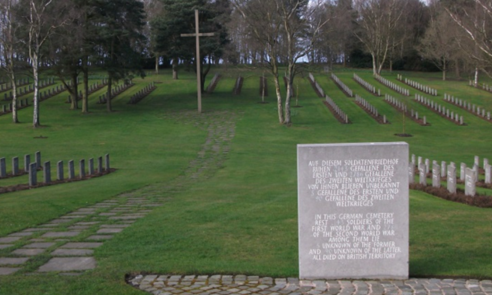 A large dedication stone leads to a wide sloping green lawn, featuring a flagstone path, leading to burial plots at Cannock Chase German War Cemetery. Rows of granite German headstones extend across the lawn.