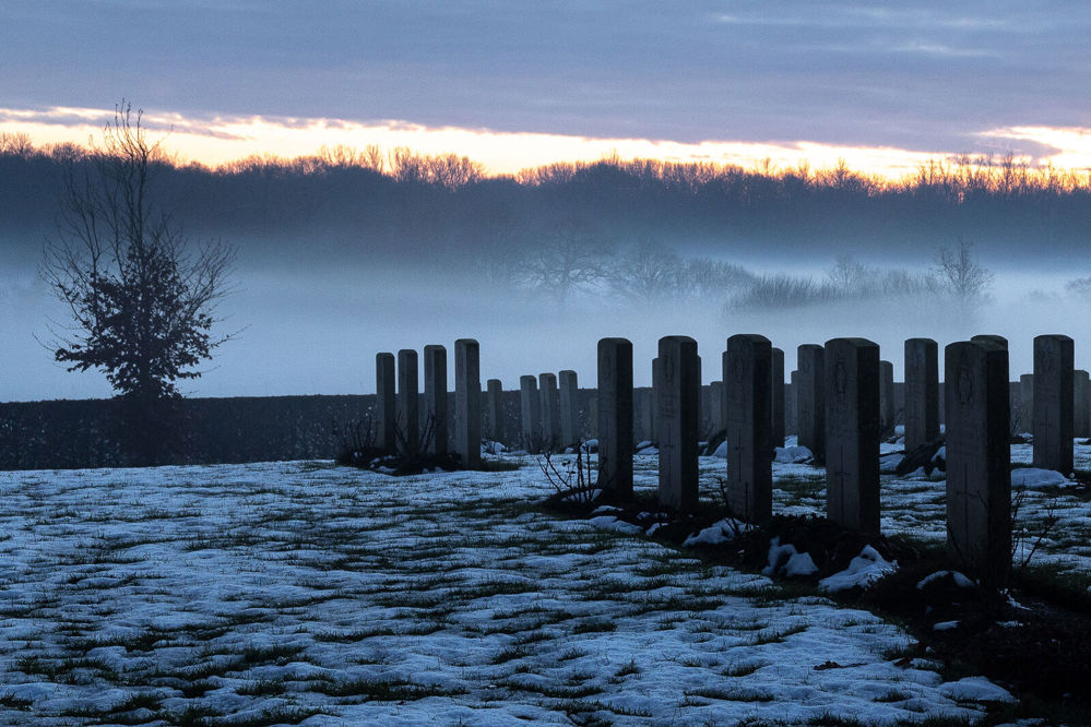 A row of headstones under a bleak Winter's morning. A thin, patchy layer of snow blankets the ground.