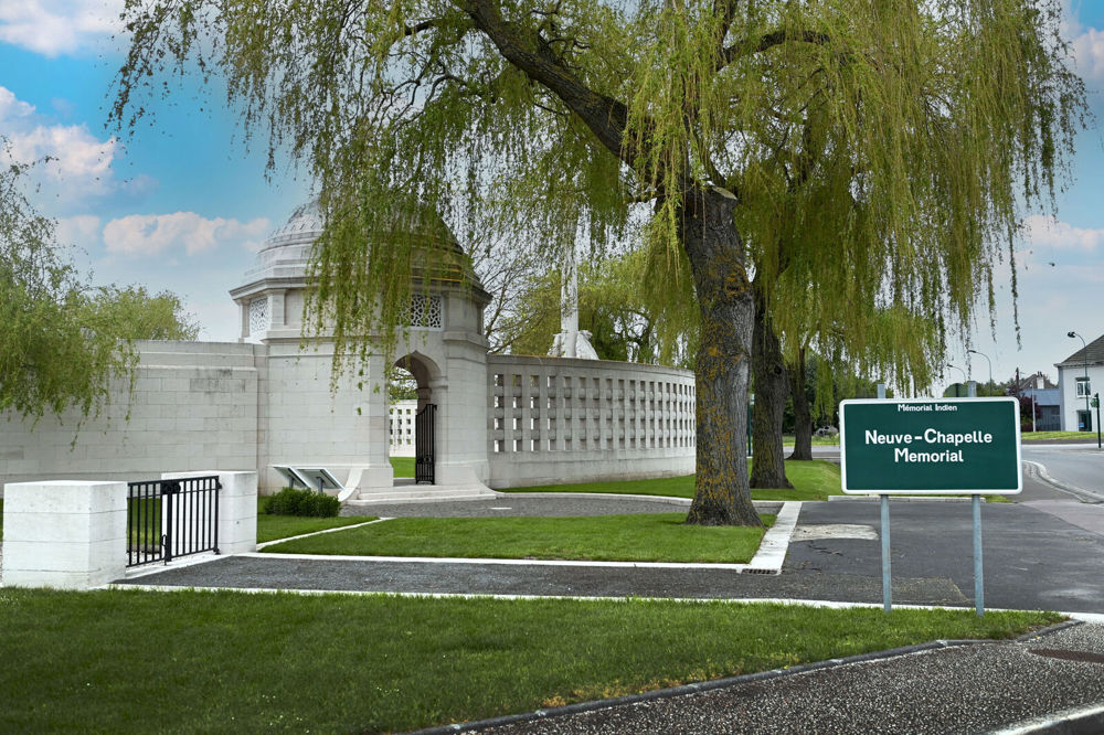 View of the Neuve-Chapelle Memorial exteriror, showing entry pavilion underneath a row of tall willow-like trees. A green CWGC sign with "Neuve-Chapelle Memorial" in white lettering is visible in the foreground.
