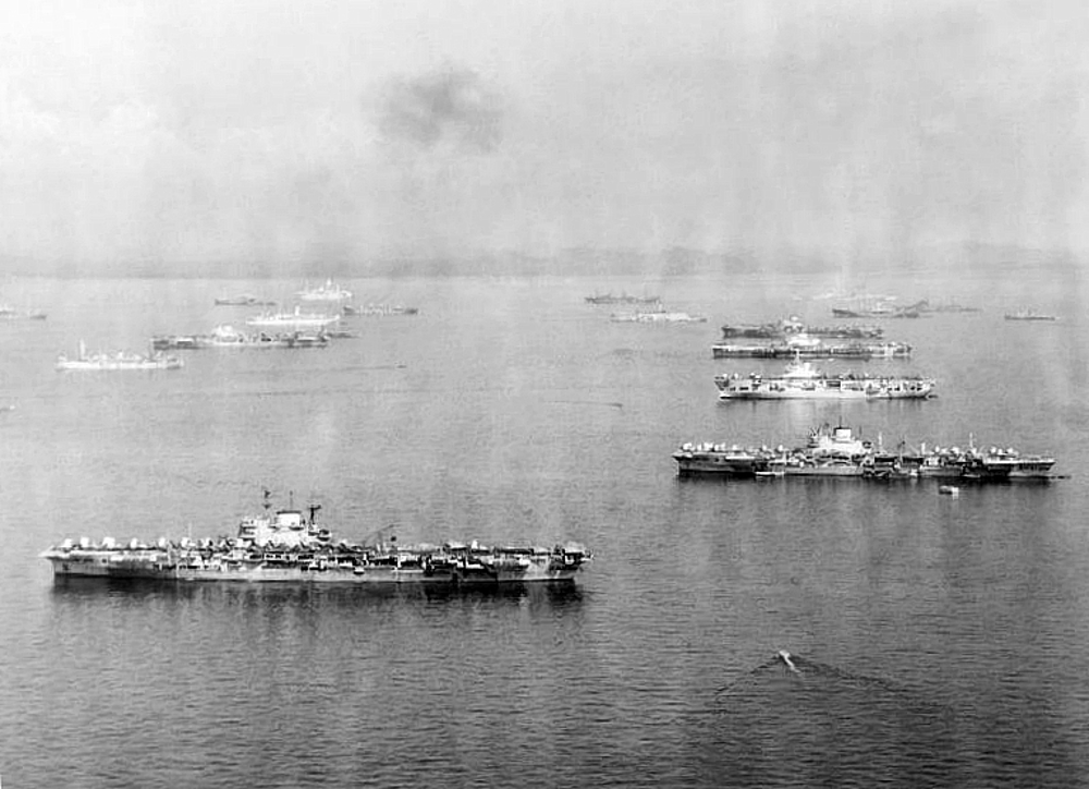 Aircraft carriers of the British Pacific Fleet at anchor in line formation. Various support and escort vessels are visible floating on the ocean with the carrier fleet. A large plume of black smoke is visible in the centre of the secene.
