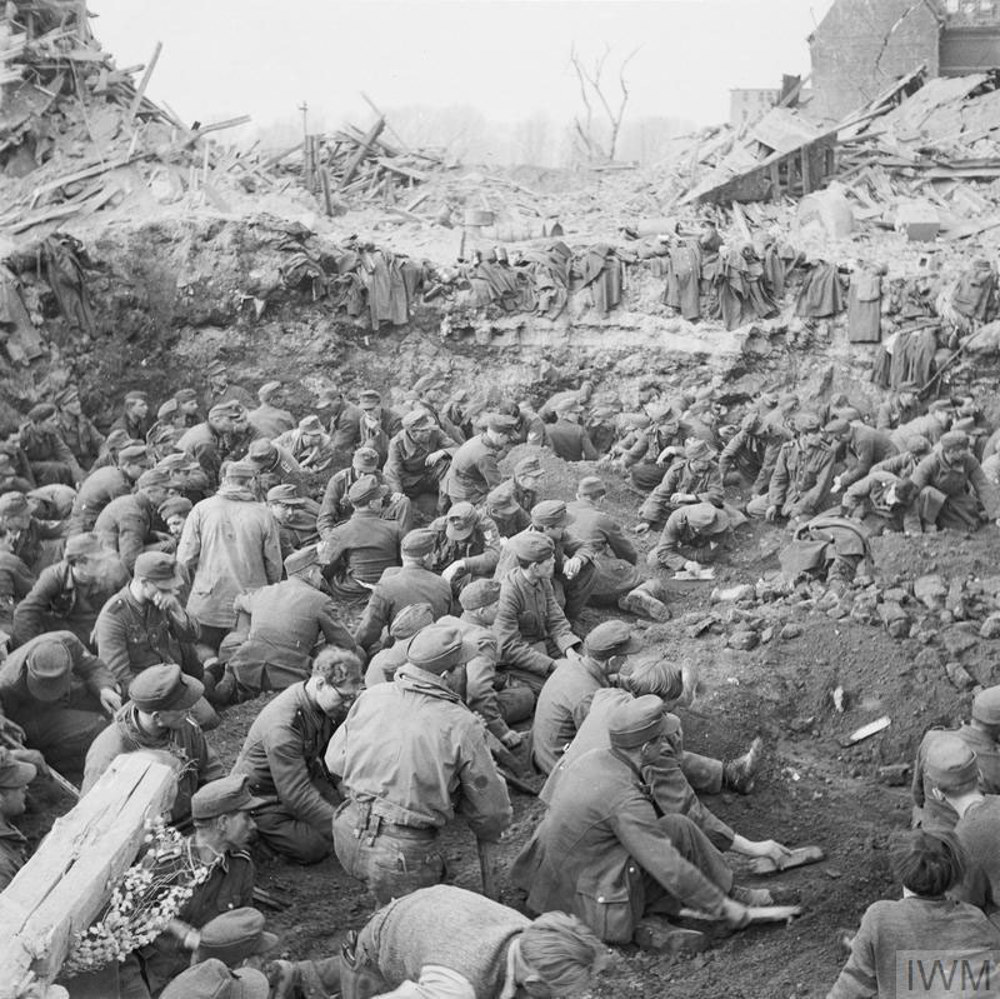 A large group of German Wehrmacht Prisoners of War site in the bombed out ruins of a city after being captured by the Allies. The soldiers are a mix of ages, in Wehrmacht uniforms. In the background are shattered buildings and rubble. The POWs are sat in a large shell crater.